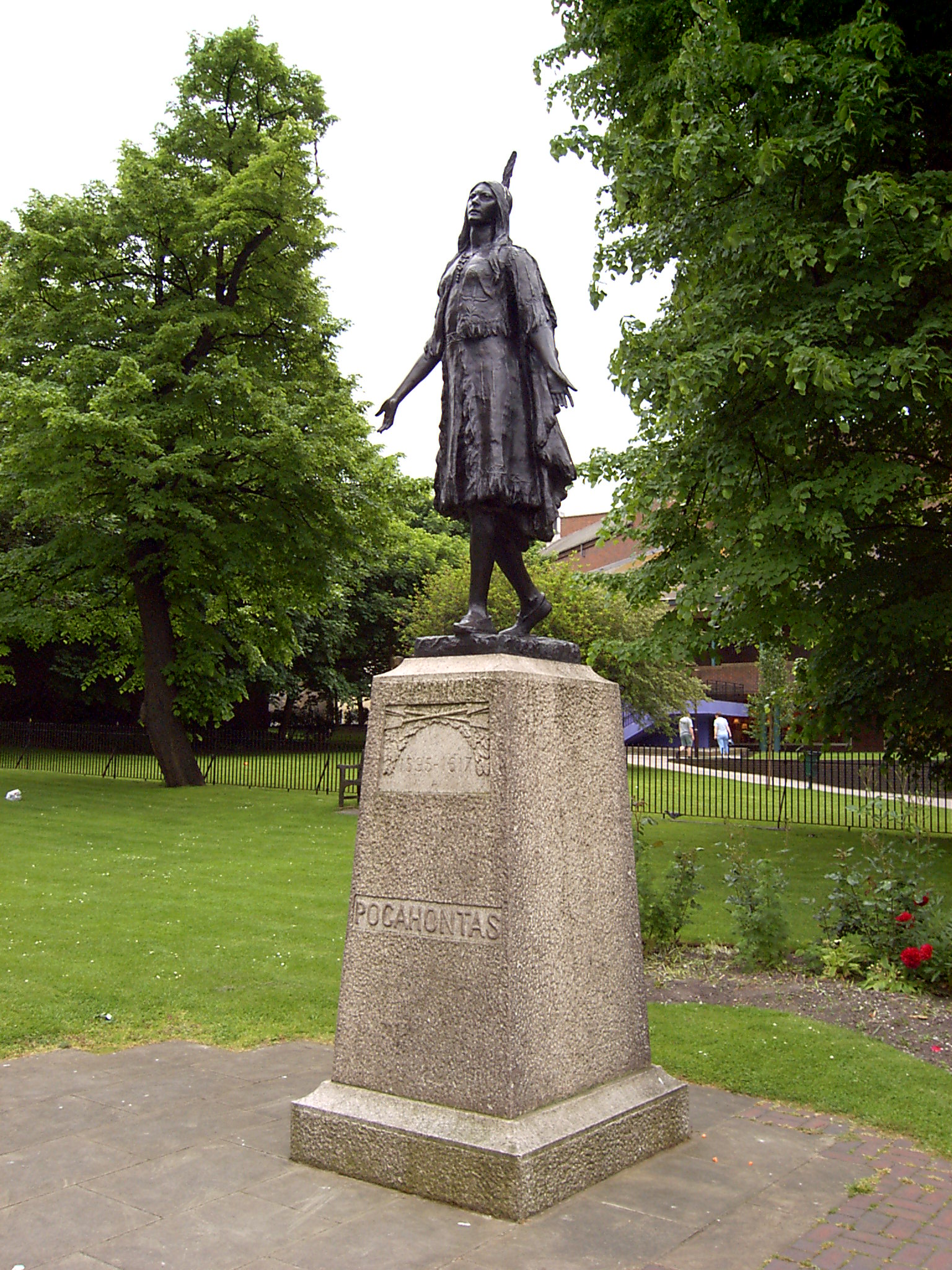Statue of Pocahontas on a stone plinth with concrete around, grass and trees behind. She wears a dress and a feather in her hair. She holds her arms down, but with her hands raised slightly as if in greeting.