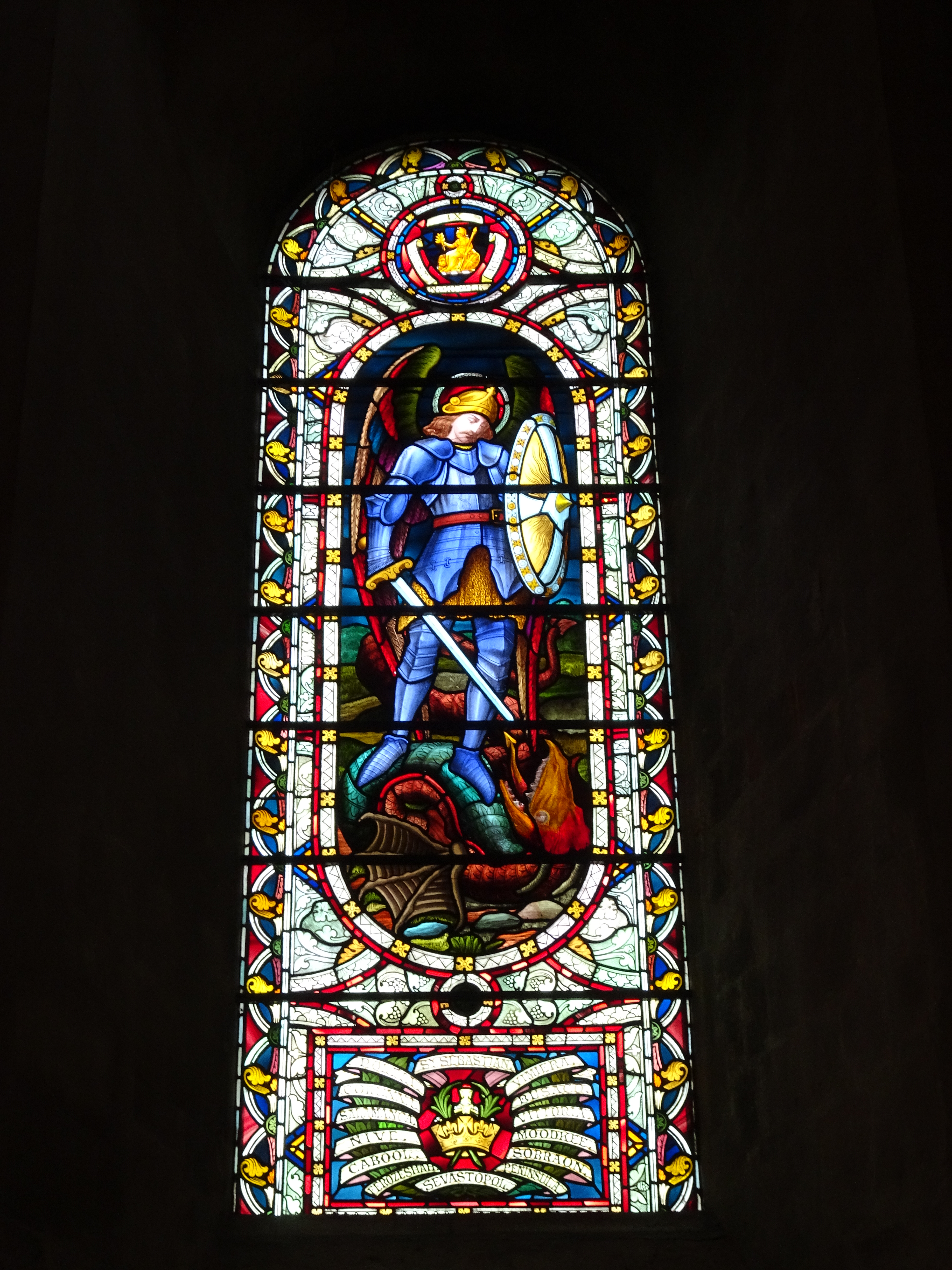Stained glass window in Norwich Anglican Cathedral showing St Michael in blue armour, with sword and shield, standing on the dragon.